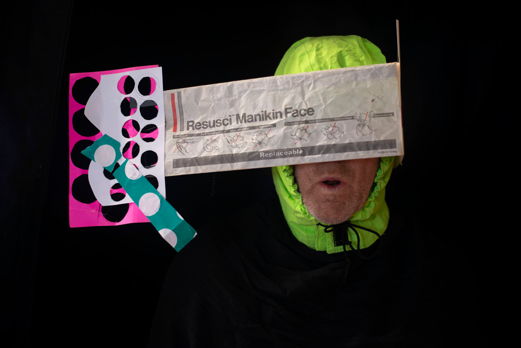 On a black background appears with a head wearing a florescent yellow hood, and the face is largely covered by a paper form that stretches to the left of the head.