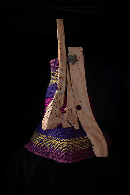 On a black background sits a vertical, conical form consisting of woven fabric in yellows, blues and purples, fronted by some random wood pieces.