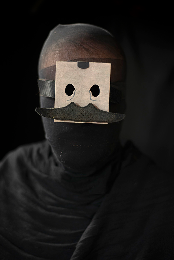 A shadowy figure wearing a small square light color mask on a black background.