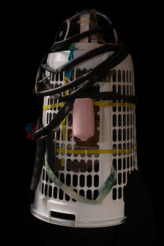 In front of a black background sits a sculpture of an inverted tall white plastic clothes basket festooned with a number of items.