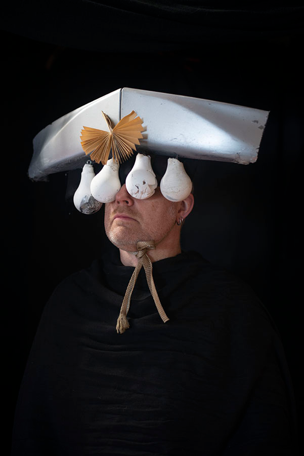 A male face turned to the left wearing a headpiece mask made of lightbulbs hanging from a Chevron shaped piece of plastic that covers the eyes.