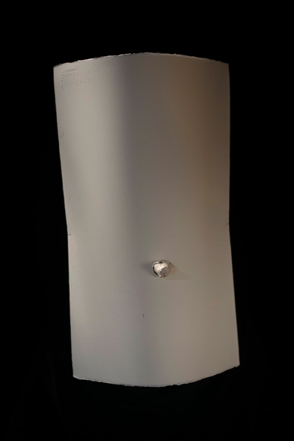 A cylindrical white vertical form with a small protrusion in its lover center sits over a black background.